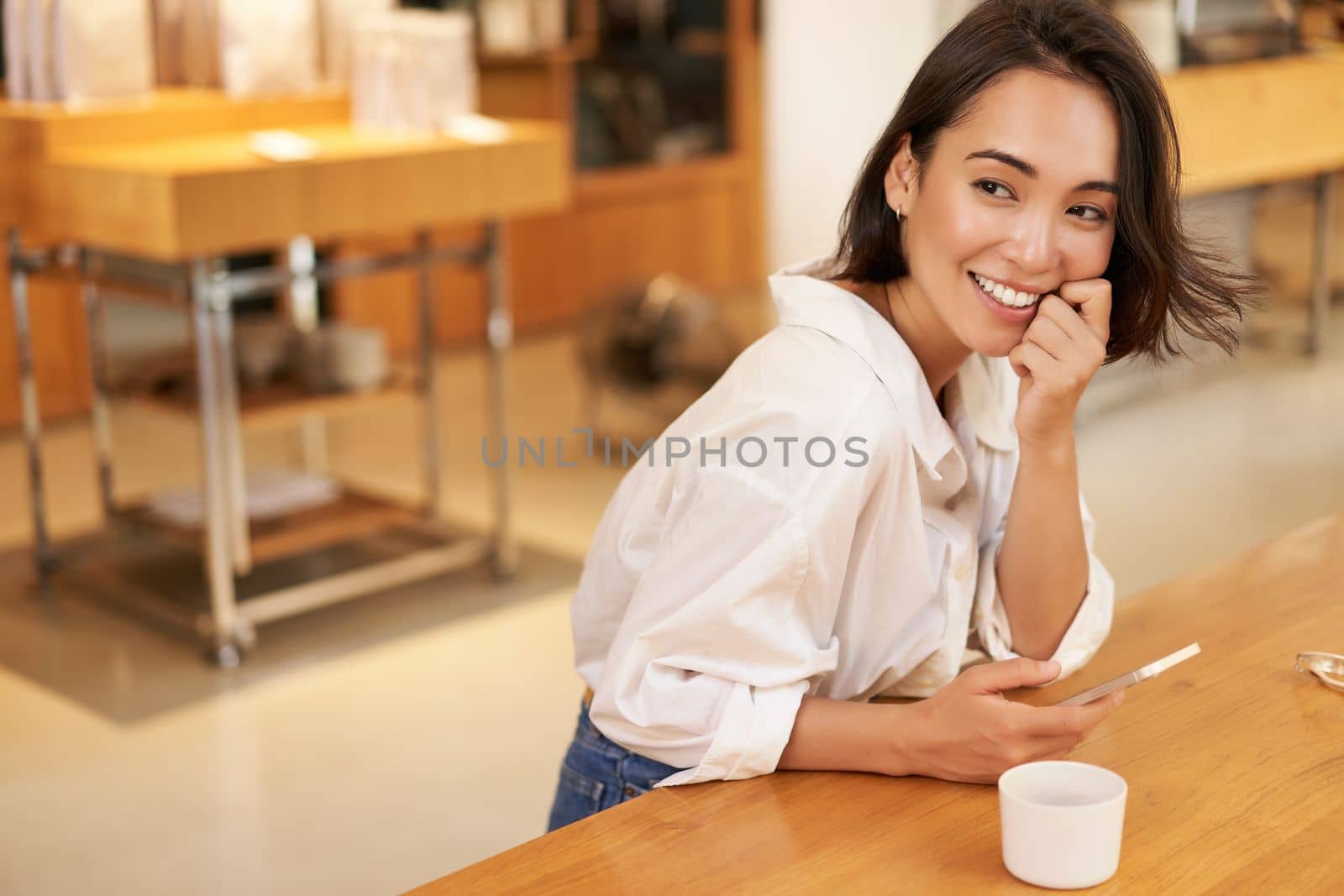 Portrait of young asian woman, sitting in cafe, holding smartphone, chatting and messaging while drinking coffee.