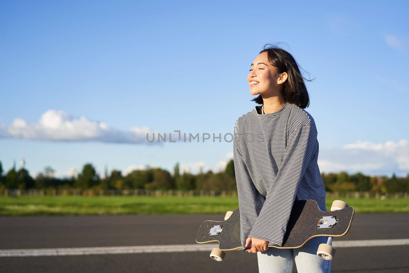 Portrait of asian woman with longboard. Korean girl skating, holding skateboard in hands, posing on road, smiling and looking aside by Benzoix