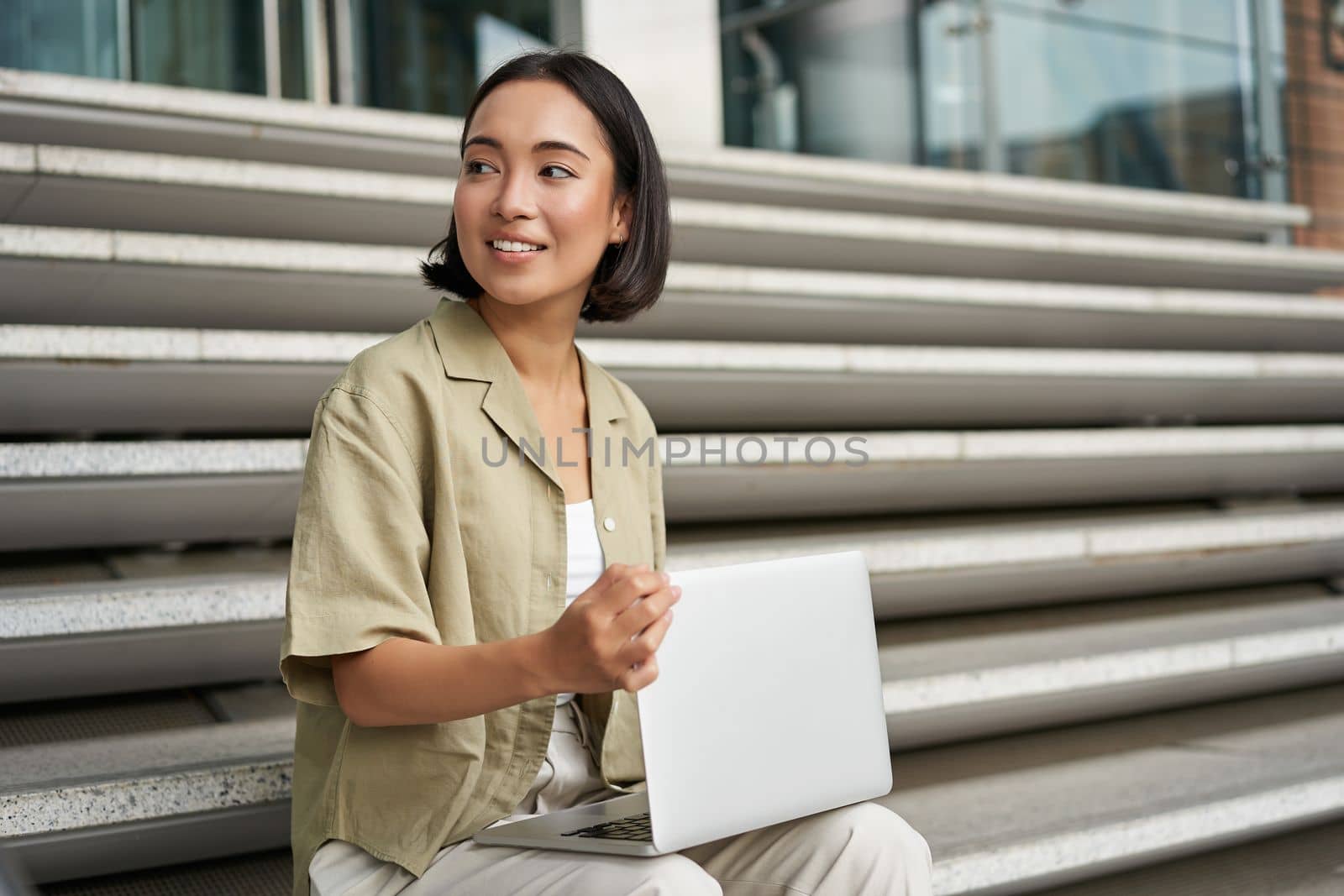 Portrait of young woman student, asian girl using laptop. Asign smiling girl, digital nomad works on her project remotely, sitting on street.