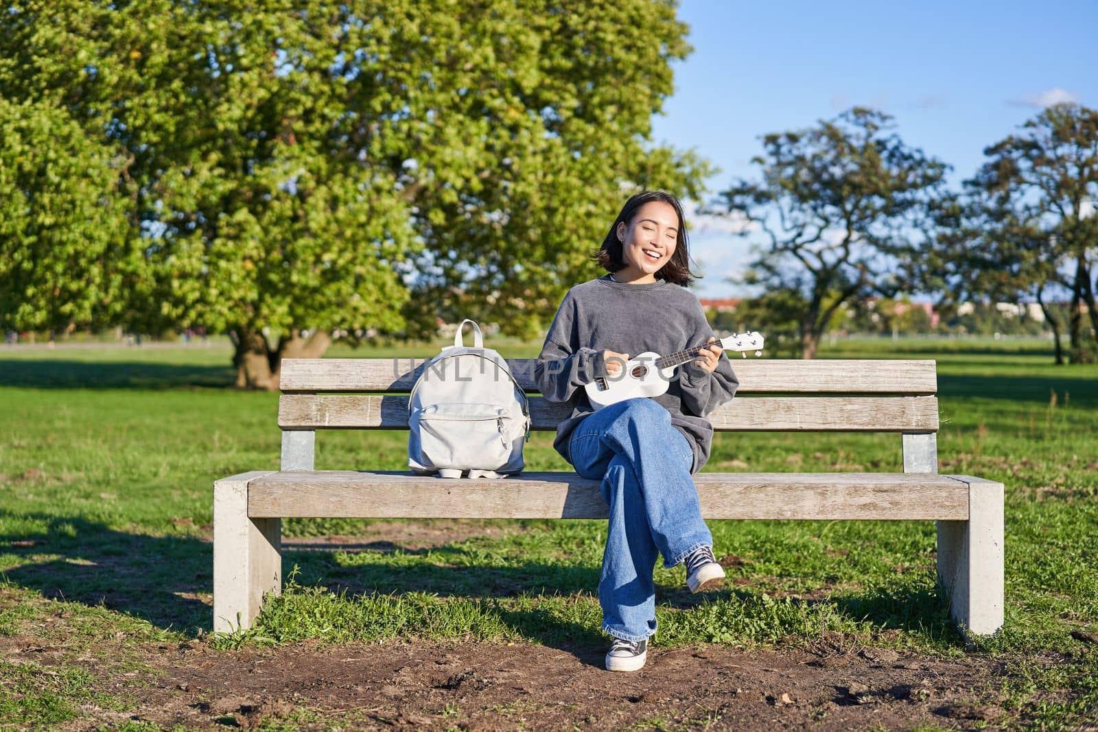 Carefree girl sits on bench in park with ukulele, plays and sings outdoors on sunny happy day.