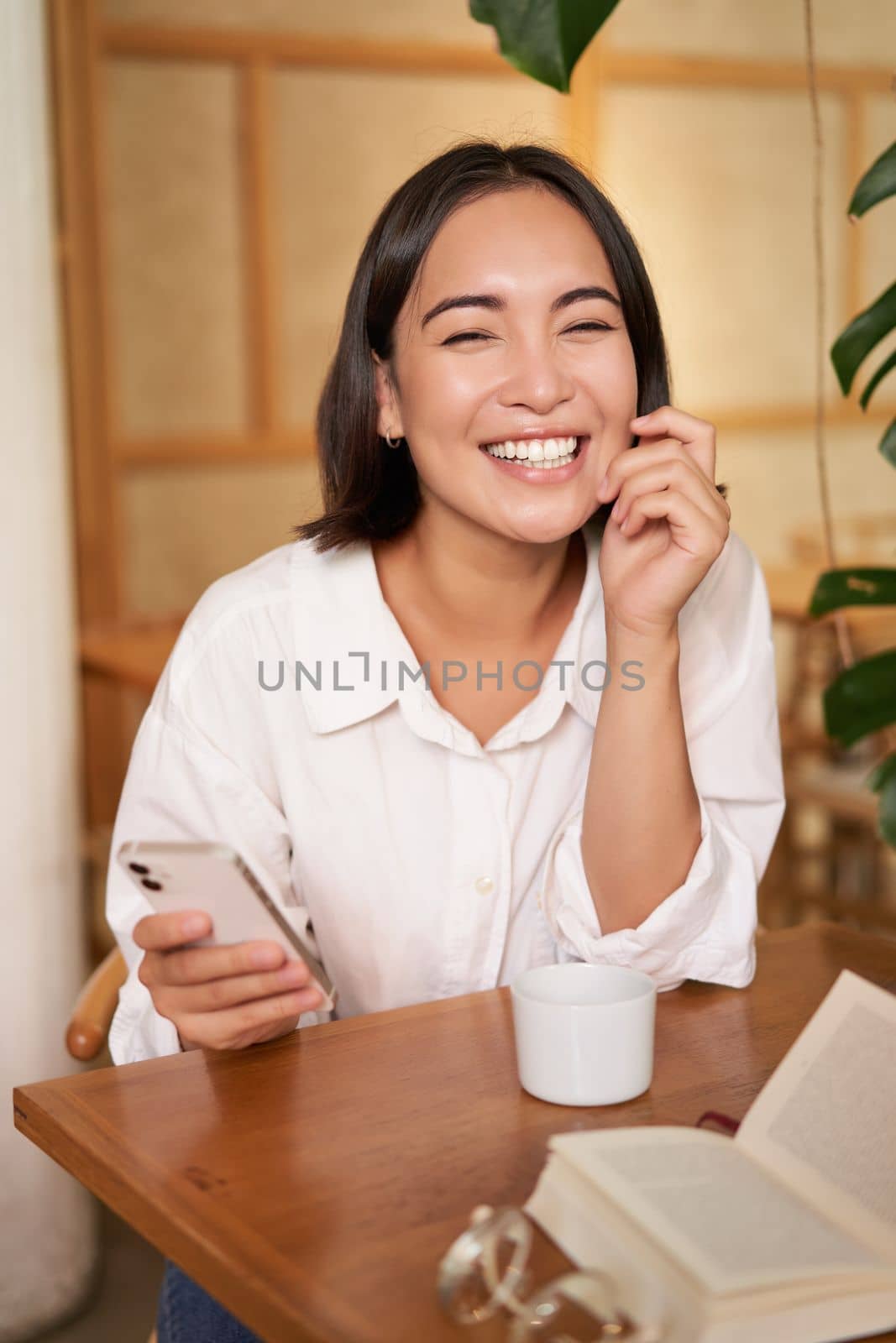 Stylish young woman in cafe with coffee, holding mobile phone, laughing and smiling. Lifestyle and people concept.