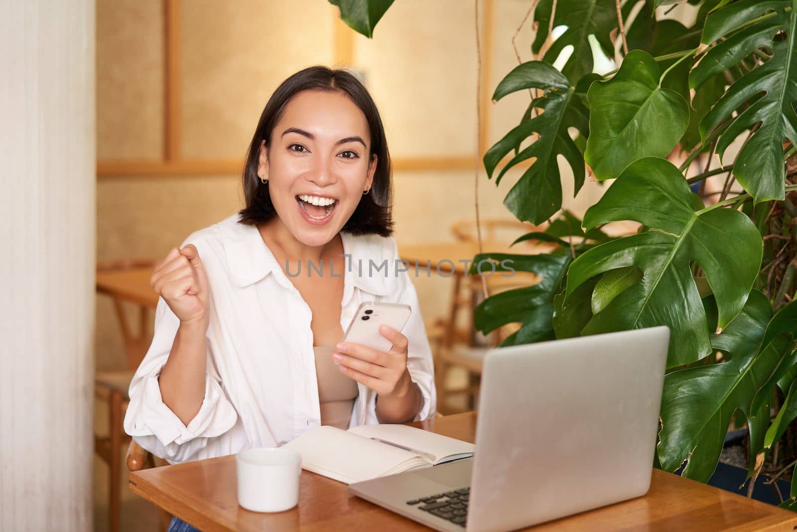 Joyful young asian woman winning, celebrating victory or achievement, receive good news on mobile phone, sitting with laptop and working by Benzoix