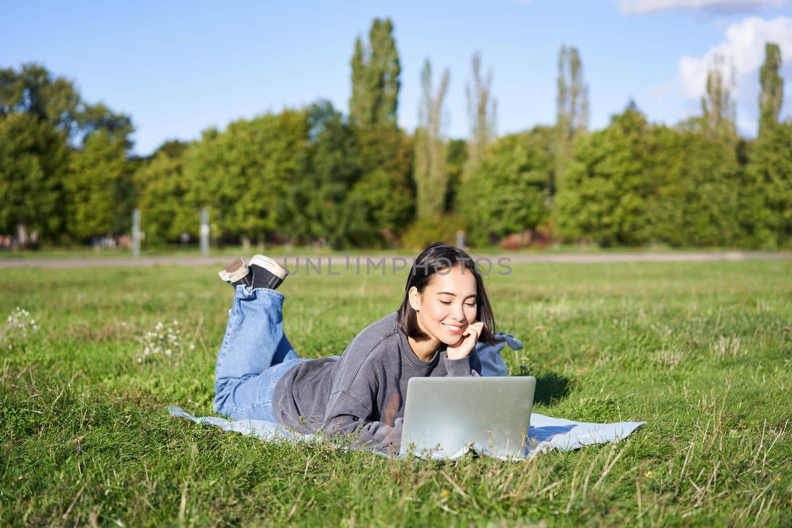 Portrait of young happy asian girl lying on blanket in park, watching videos and browsing internet on her laptop by Benzoix