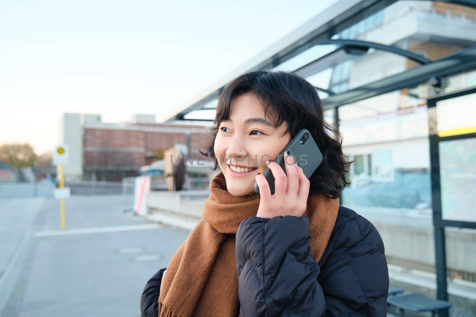 Smilling Korean girl talking on mobile phone, standing on bus stop, using smartphone, posing on road in winter, wrapped in scarf, wearing black jacket.