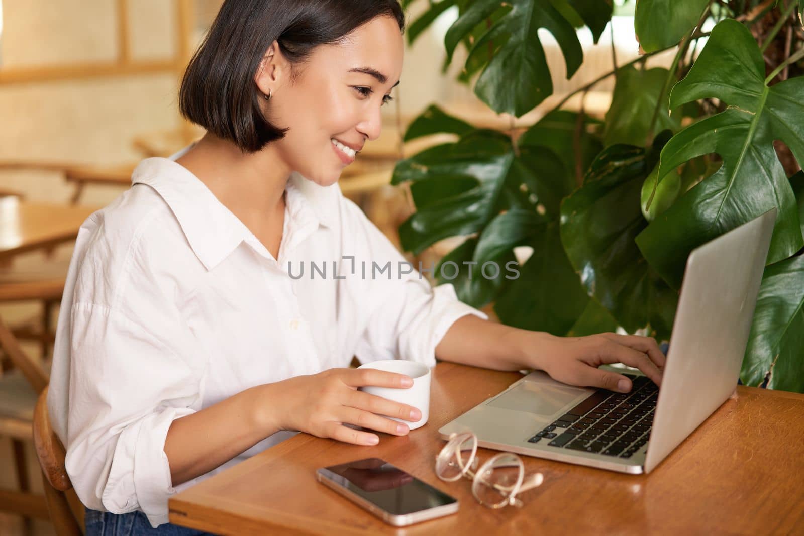 Student sitting in cafe with cup of coffee. Young asian woman working on laptop in restaurant, sitting with computer and smartphone.