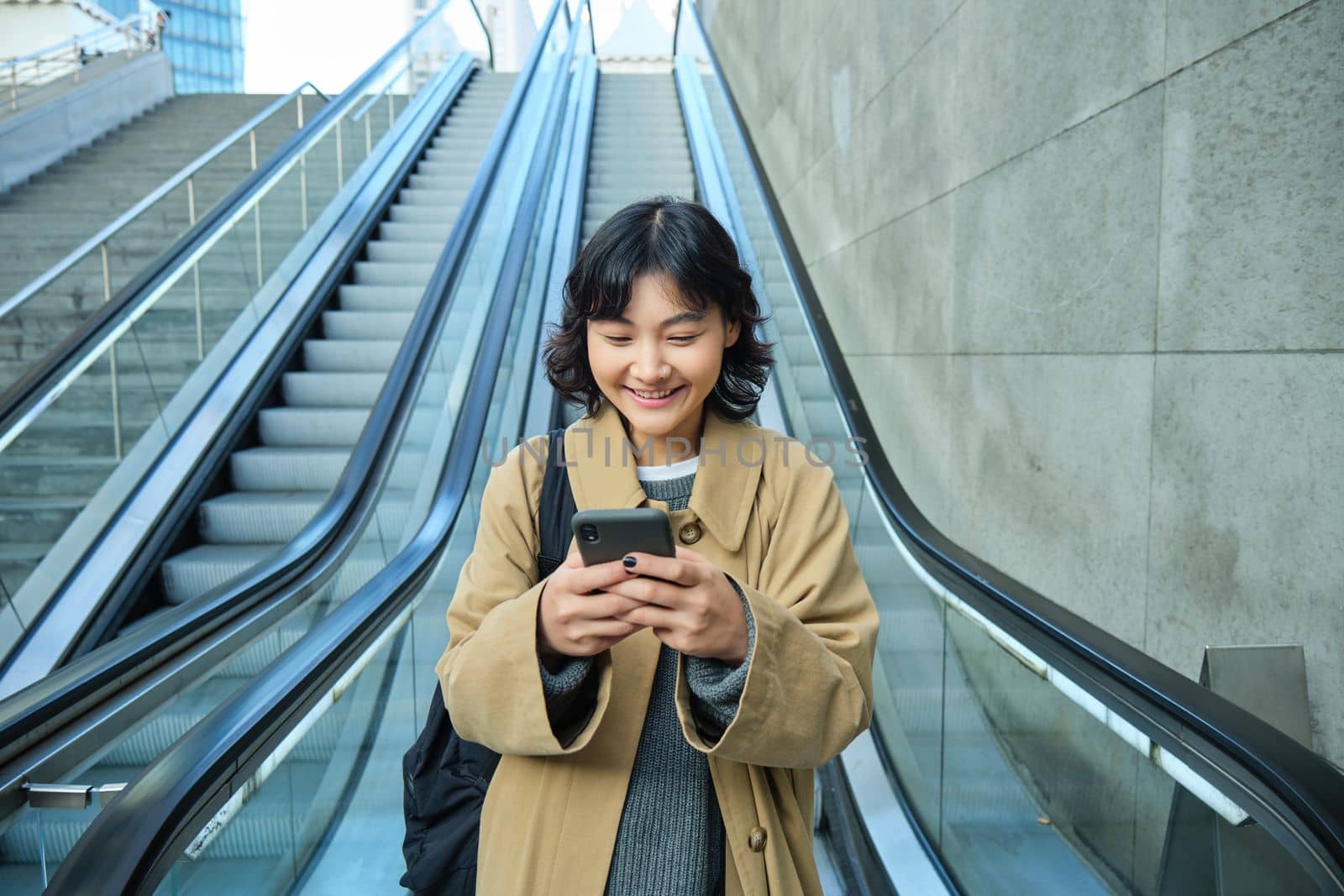 Portrait of beautiful asian girl, student goes down escalator in city, looks at mobile phone, uses telephone, map application, commutes somewhere in town by Benzoix