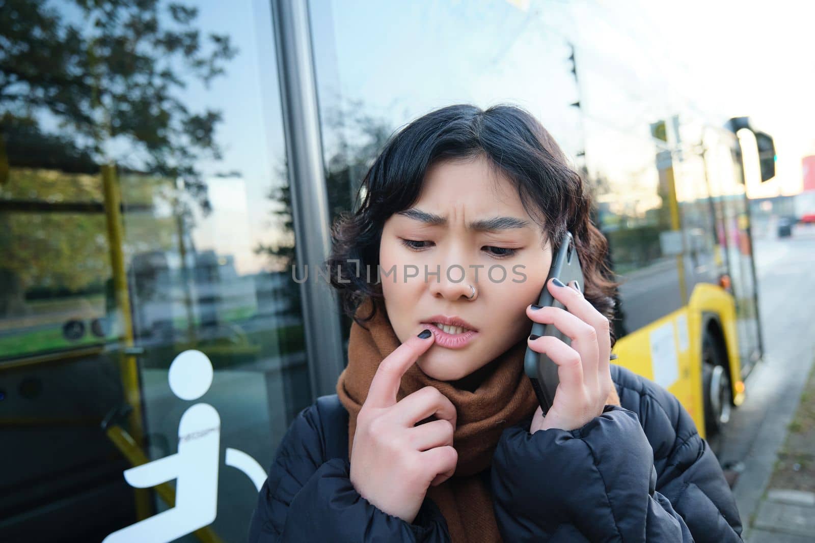 Perplexed asian girl, talks on mobile phone with concerned, thoughtful face, stands near bus stop and frowns, thinking while listening telephone conversation.