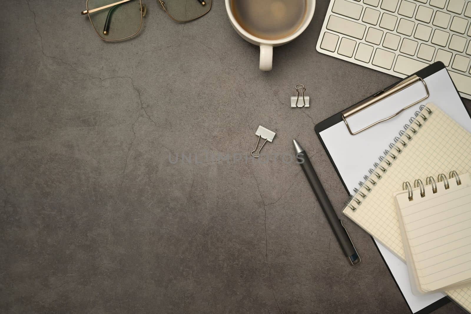 Stylish workplace with cup of coffee, keyboard, blank notepad and glasses on black slate texture background.