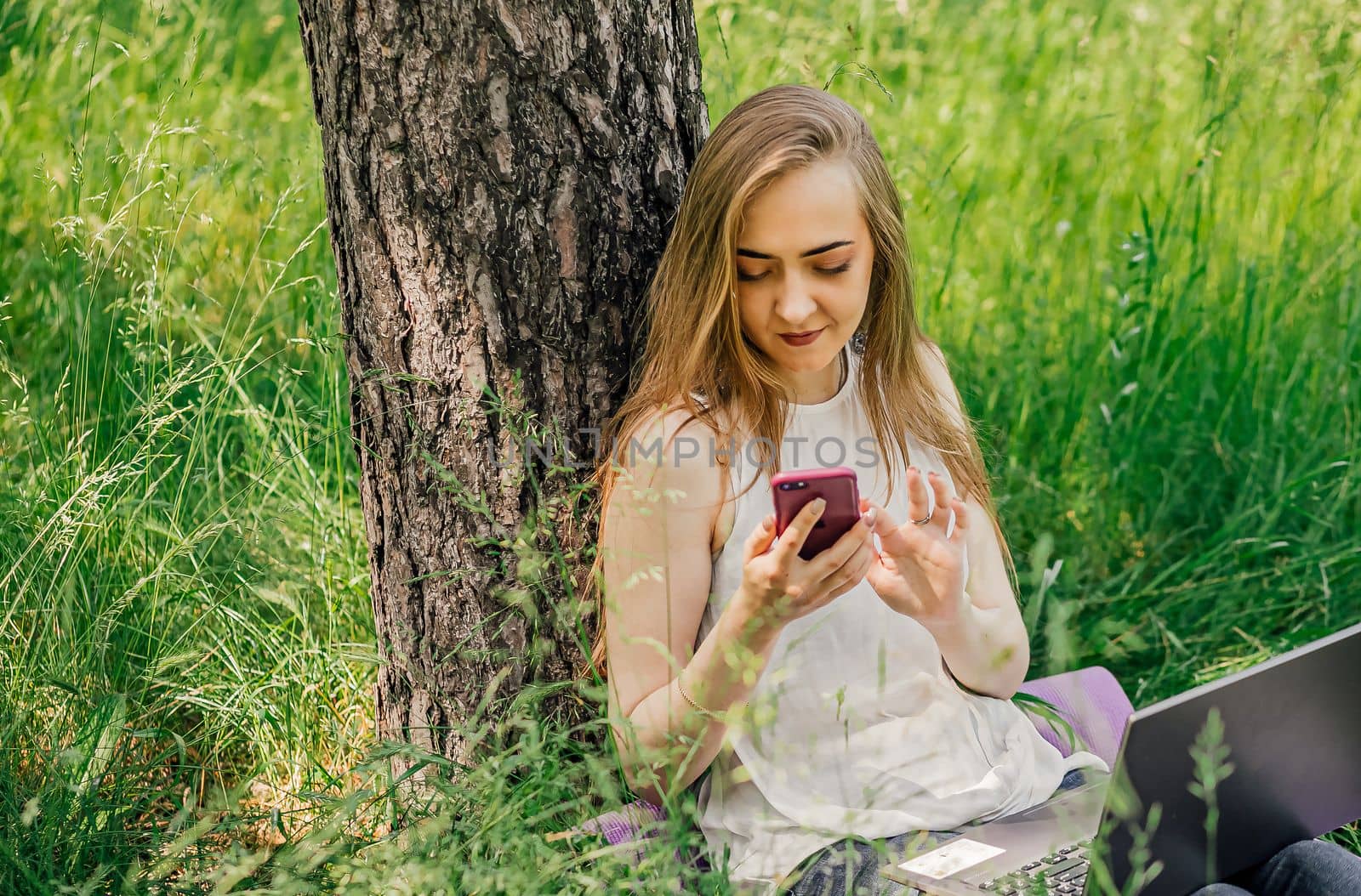 girl sits on the grass and works at a laptop. looks at the phone. freelance. selfeducation. the concept of remote learning and outdoor work