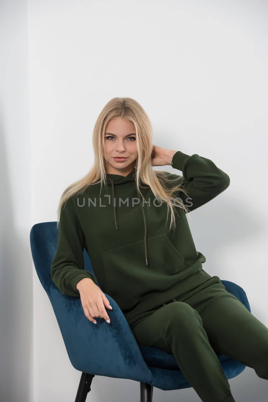 Stylish beautiful young blond woman in a green tracksuit poses near a white wall in the room. Attractive girl model posing in a blue chair. Fitness lady