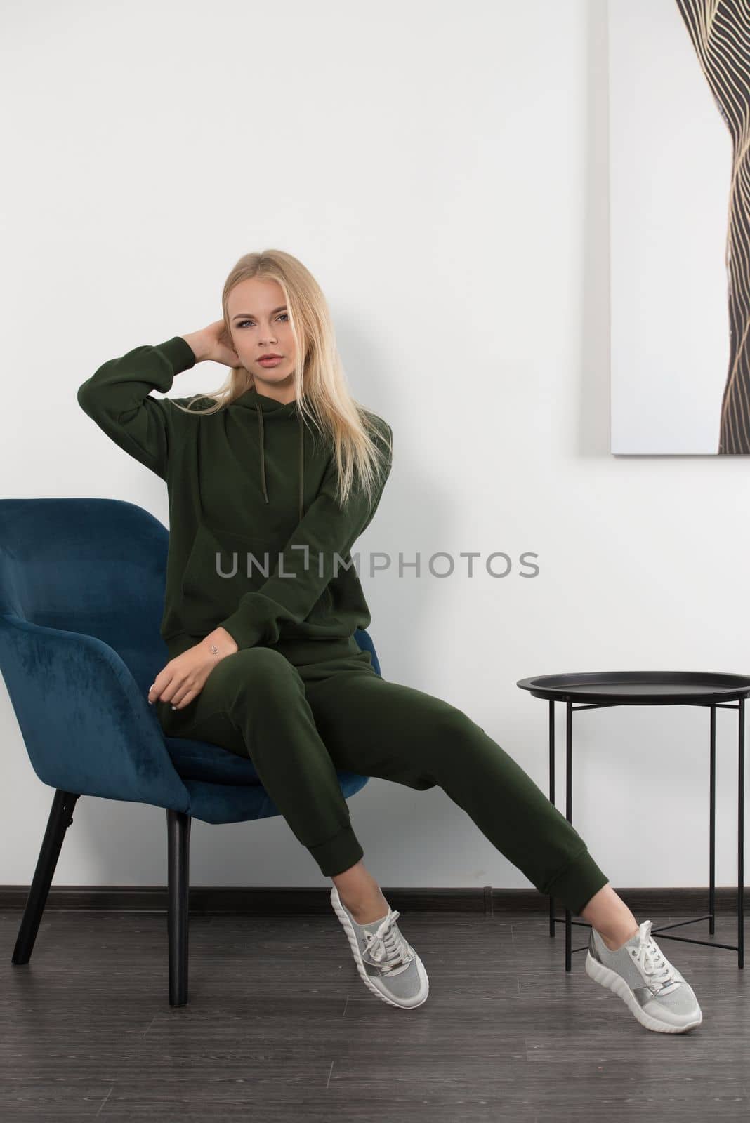 Stylish beautiful young blond woman in a green tracksuit poses near a white wall in the room. Attractive girl model posing in a blue chair. by Ashtray25