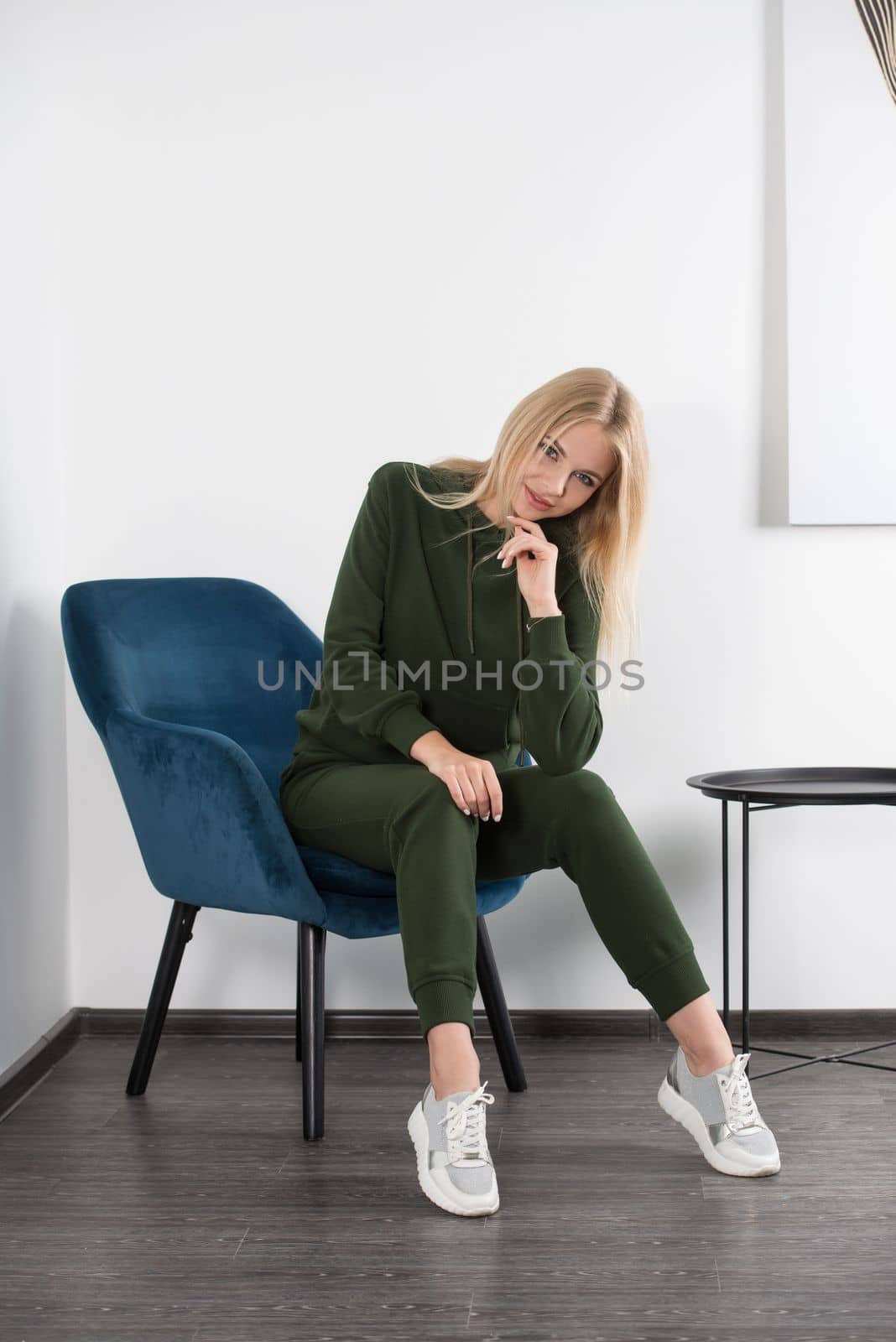 Stylish beautiful young blond woman in a green tracksuit poses near a white wall in the room. Attractive girl model posing in a blue chair. by Ashtray25