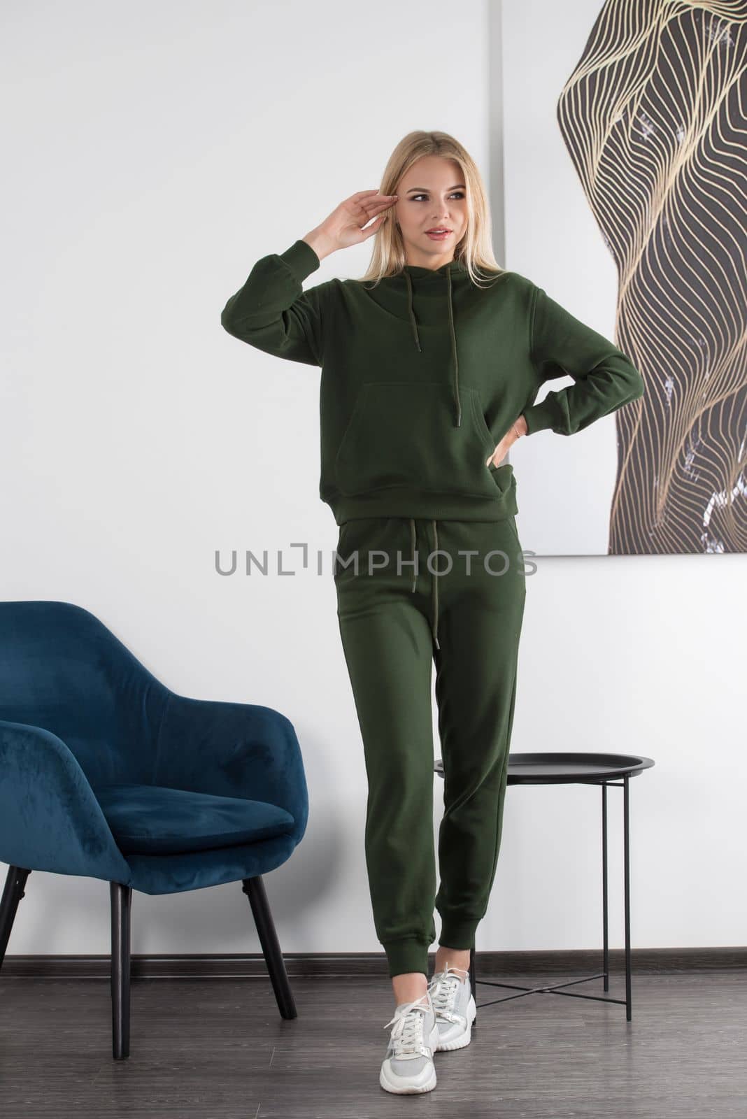 Stylish beautiful young blond woman in a green tracksuit poses near a white wall in the room. Attractive girl model posing near blue chair. by Ashtray25