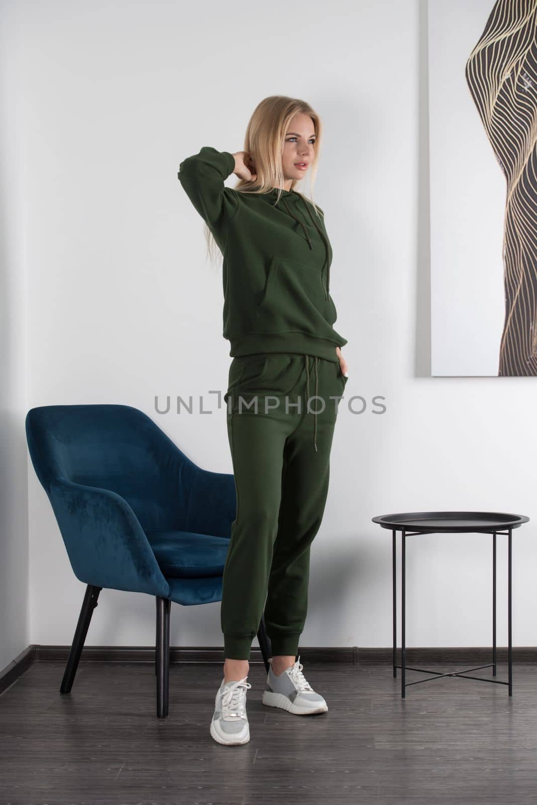 Stylish beautiful young blond woman in a green tracksuit poses near a white wall in the room. Attractive girl model posing near blue chair. Fitness lady