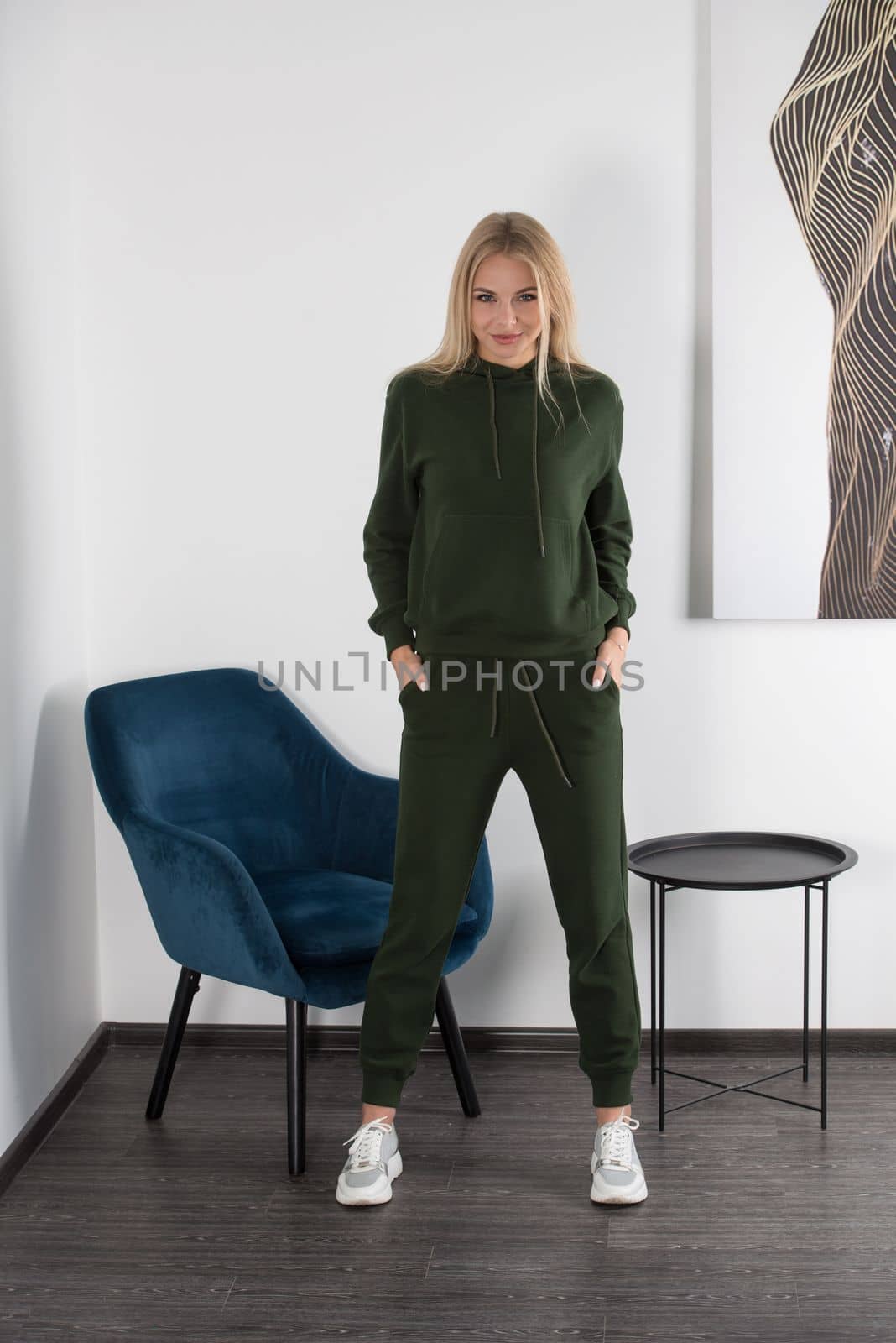 Stylish beautiful young blond woman in a green tracksuit poses near a white wall in the room. Attractive girl model posing near blue chair. by Ashtray25