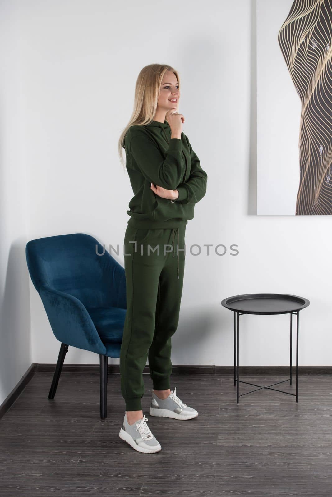 Stylish beautiful young blond woman in a green tracksuit poses near a white wall in the room. Attractive girl model posing near blue chair. Fitness lady