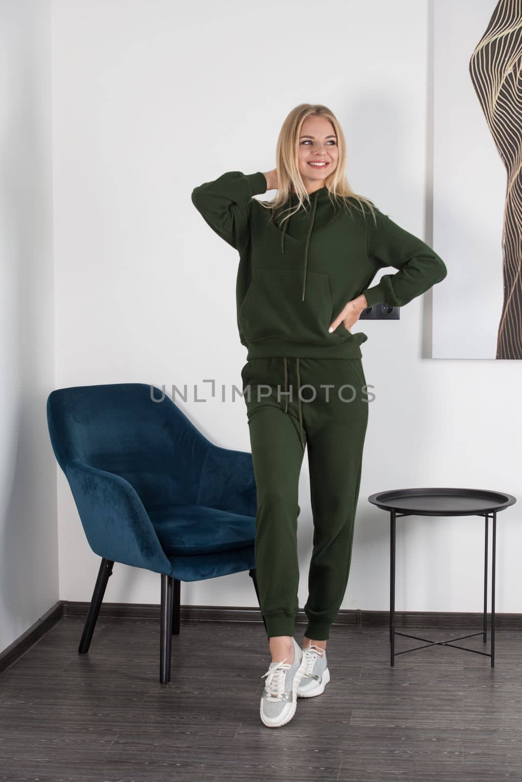 Stylish beautiful young blond woman in a green tracksuit poses near a white wall in the room. Attractive girl model posing near blue chair. by Ashtray25