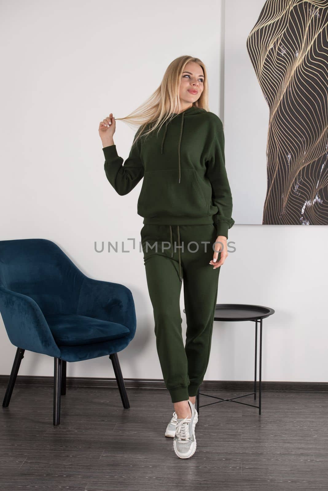 Stylish beautiful young blond woman in a green tracksuit poses near a white wall in the room. Attractive girl model posing near blue chair. Fitness lady