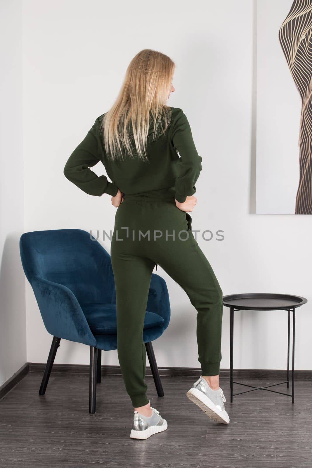 Stylish beautiful young blond woman in a green tracksuit poses near a white wall in the room. Attractive girl model posing near blue chair. by Ashtray25