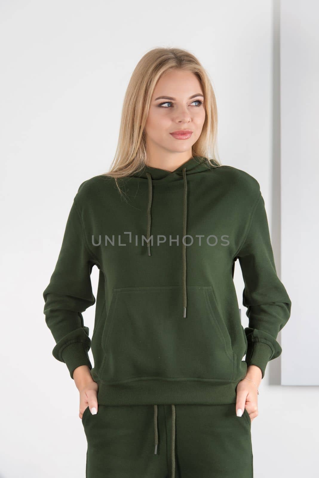 Stylish beautiful young blond woman in a green tracksuit poses near a white wall in the room. by Ashtray25