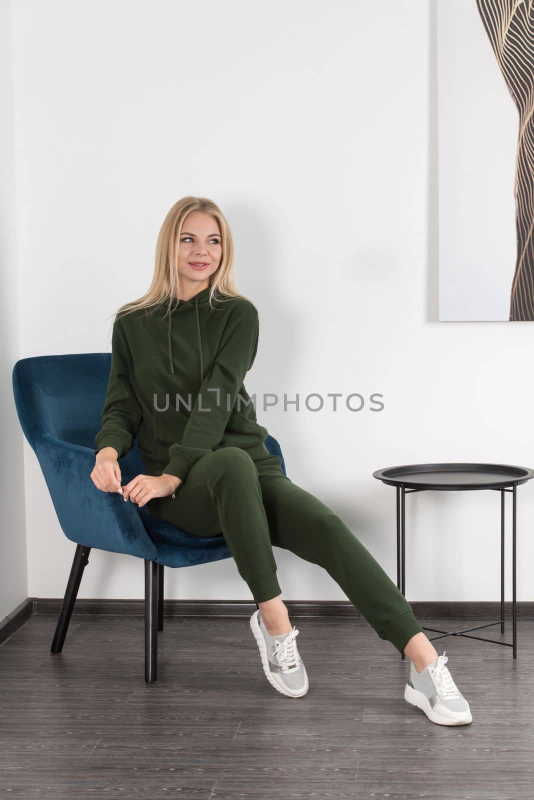 Stylish beautiful young blond woman in a green tracksuit poses near a white wall in the room. Attractive girl model posing in a blue chair. by Ashtray25