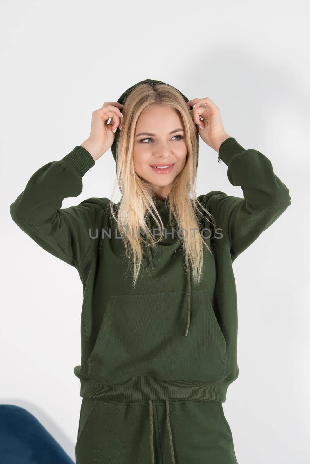 Stylish beautiful young blond woman in a green tracksuit poses near a white wall in the room. Fitness lady