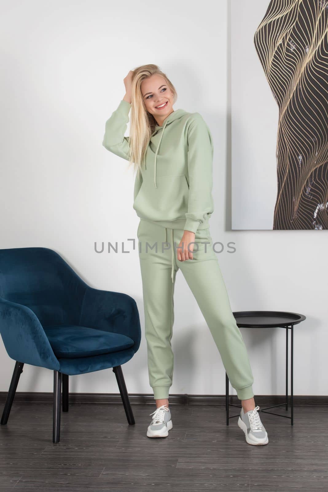 Stylish beautiful young blond woman in a light green tracksuit poses near a white wall in the room. Attractive girl model posing near blue chair. by Ashtray25