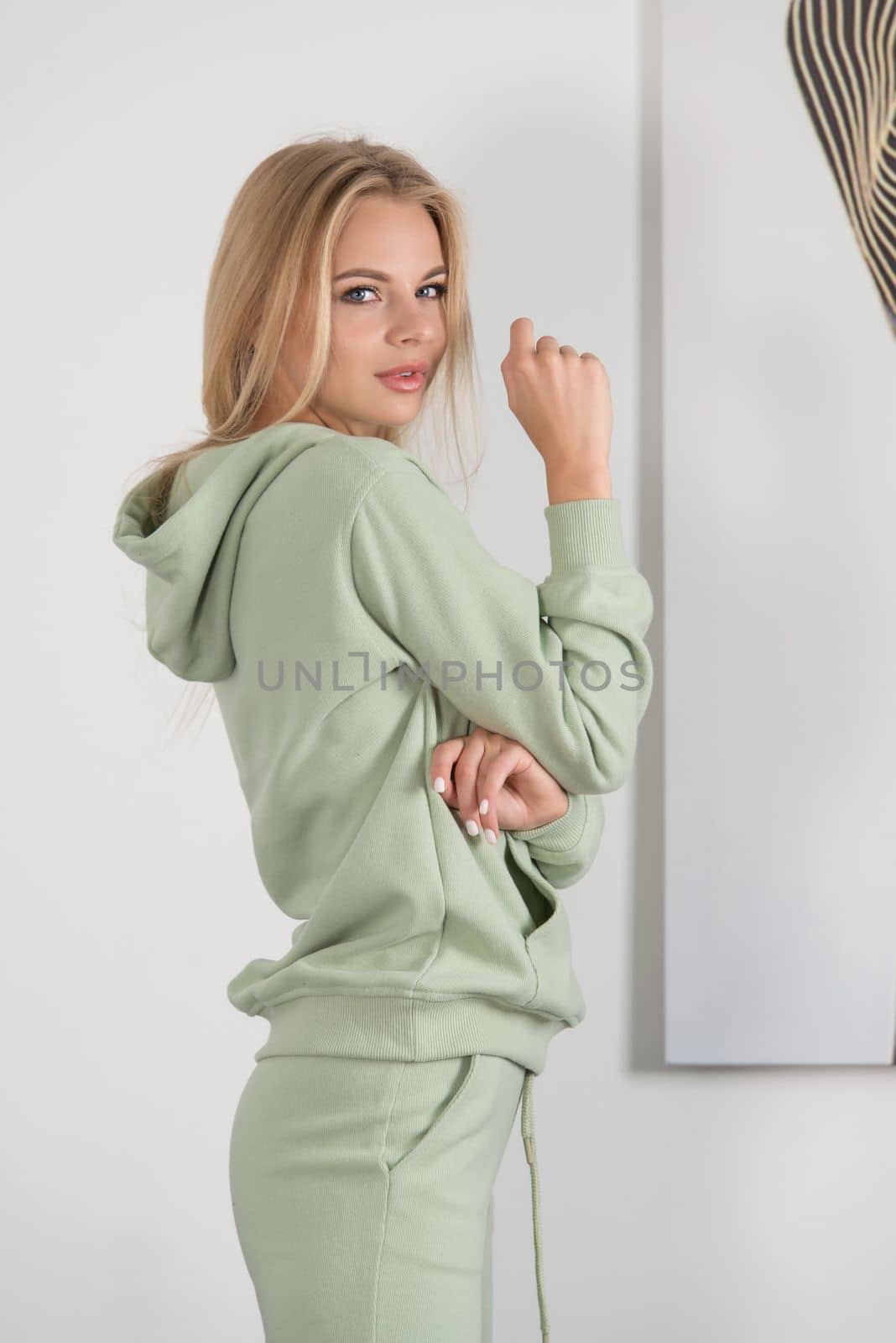 Stylish beautiful young blond woman in a green tracksuit poses near a white wall in the room. by Ashtray25