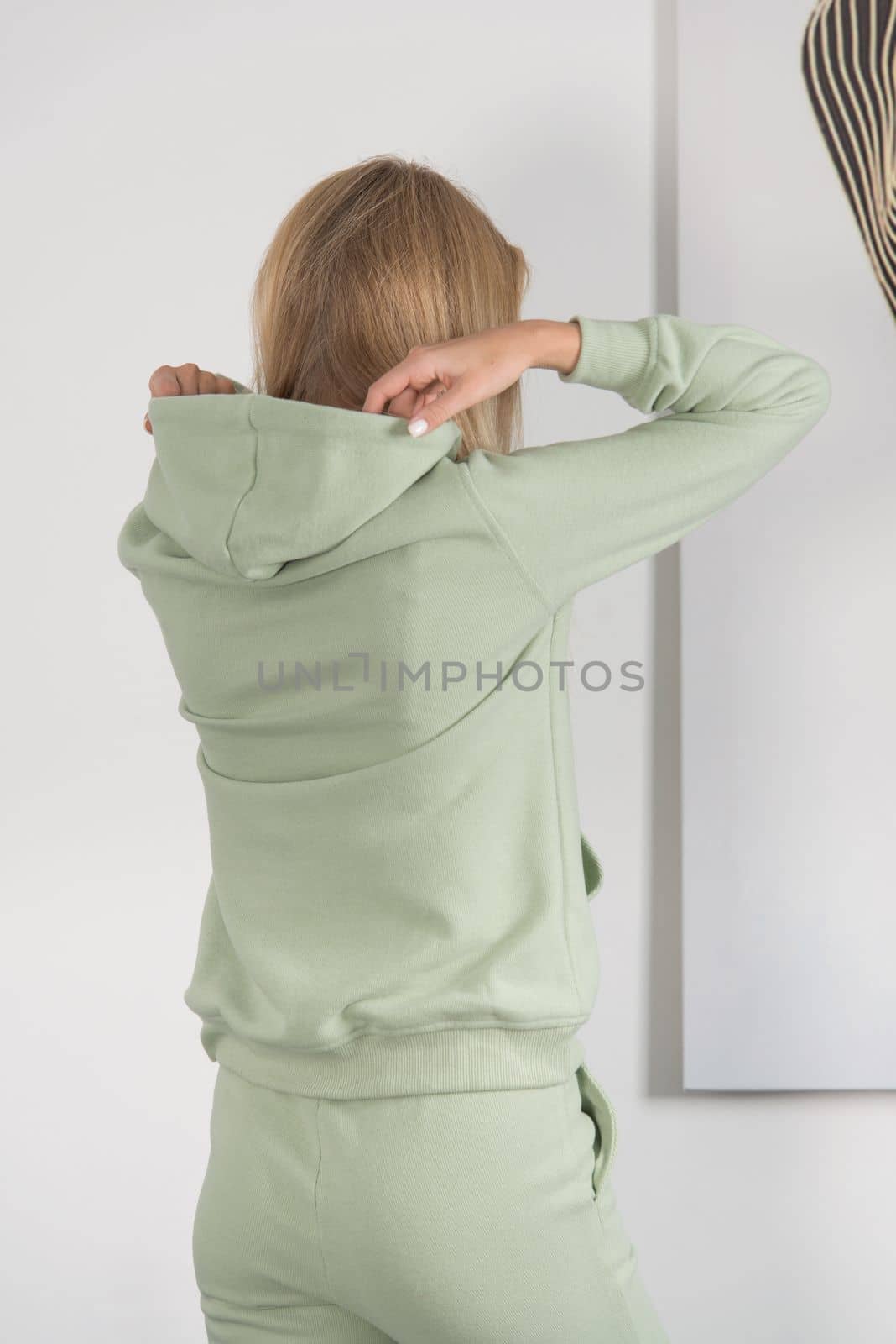 Stylish beautiful young blond woman in a green tracksuit poses near a white wall in the room. Fitness lady