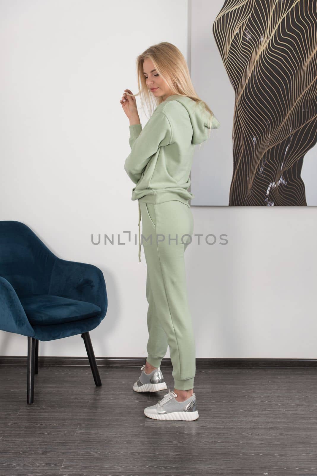 Stylish beautiful young blond woman in a light green tracksuit poses near a white wall in the room. Attractive girl model posing near blue chair. by Ashtray25