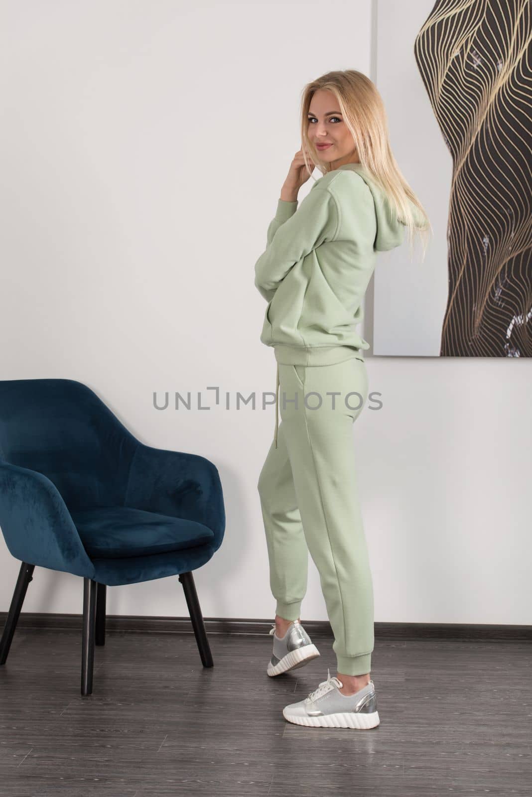 Stylish beautiful young blond woman in a light green tracksuit poses near a white wall in the room. Attractive girl model posing near blue chair. by Ashtray25