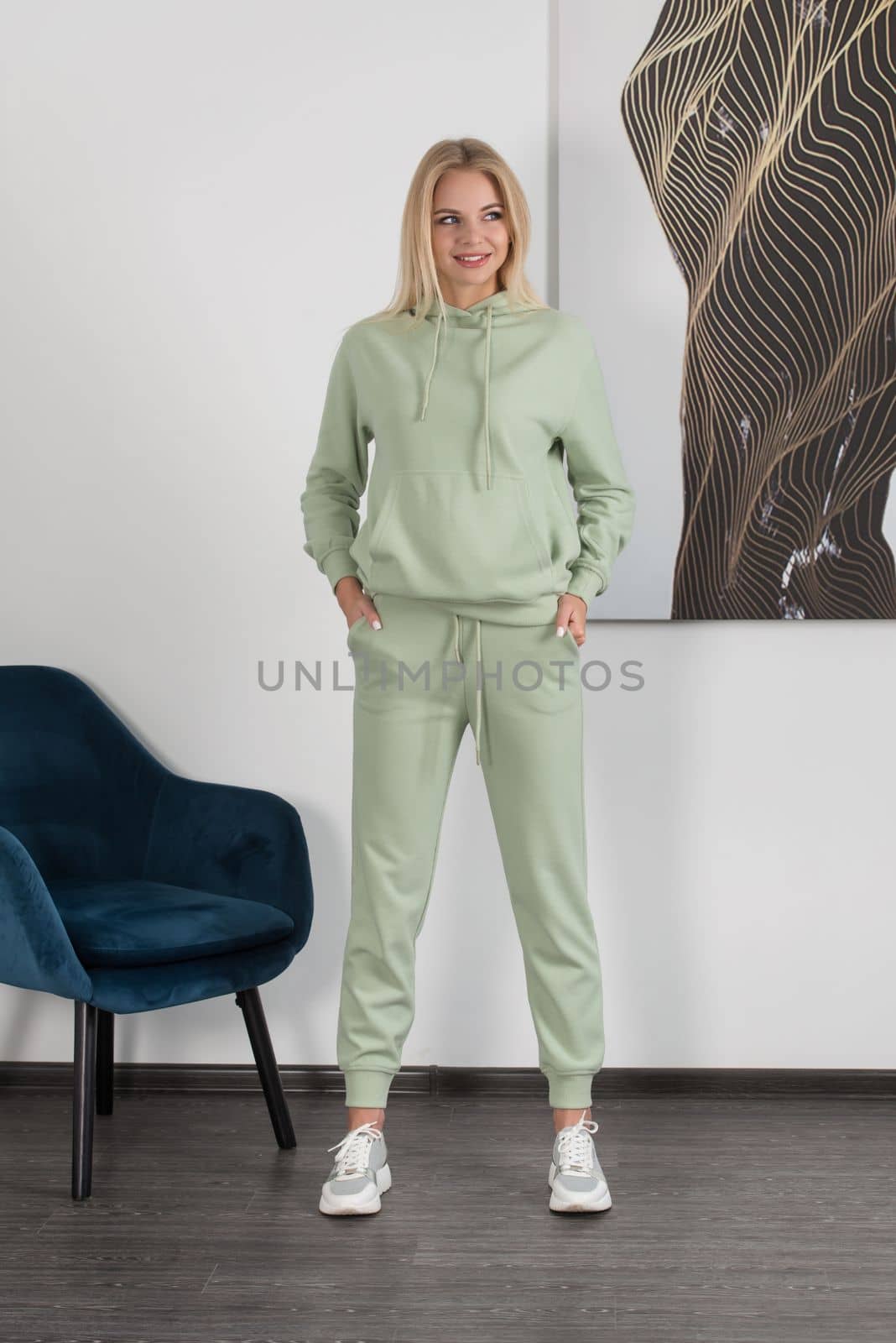 Stylish beautiful young blond woman in a light green tracksuit poses near a white wall in the room. Attractive girl model posing near blue chair. Fitness lady