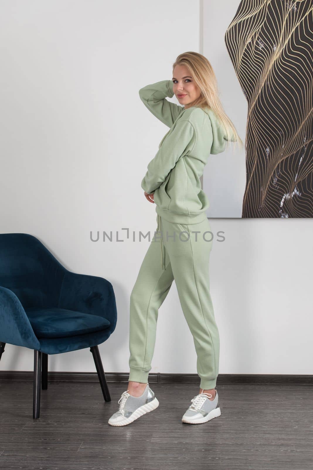 Stylish beautiful young blond woman in a light green tracksuit poses near a white wall in the room. Attractive girl model posing near blue chair. by Ashtray25