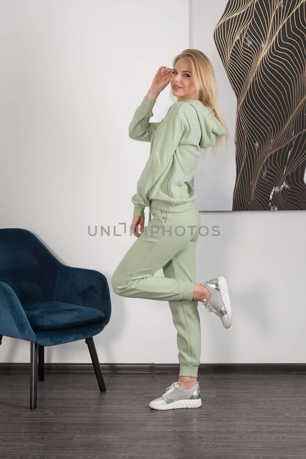 Stylish beautiful young blond woman in a light green tracksuit poses near a white wall in the room. Attractive girl model posing near blue chair. Fitness lady