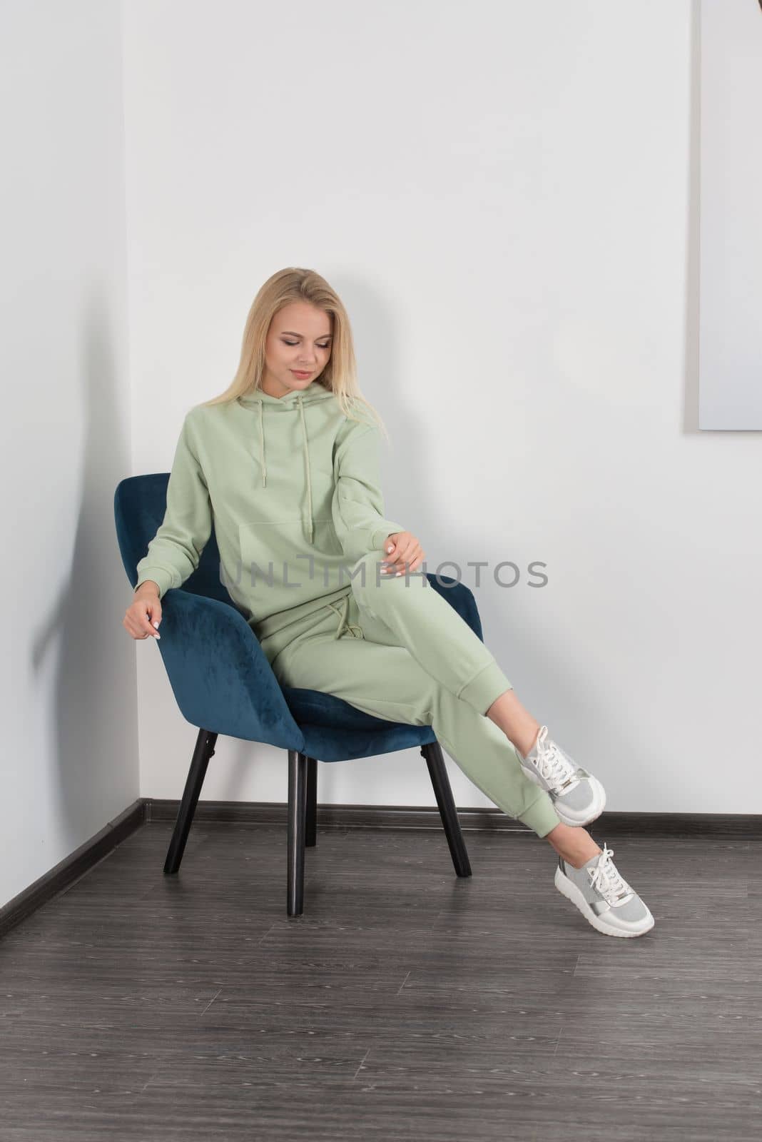 Stylish beautiful young blond woman in a light green tracksuit poses near a white wall in the room. Attractive girl model posing in a blue chair. Fitness lady