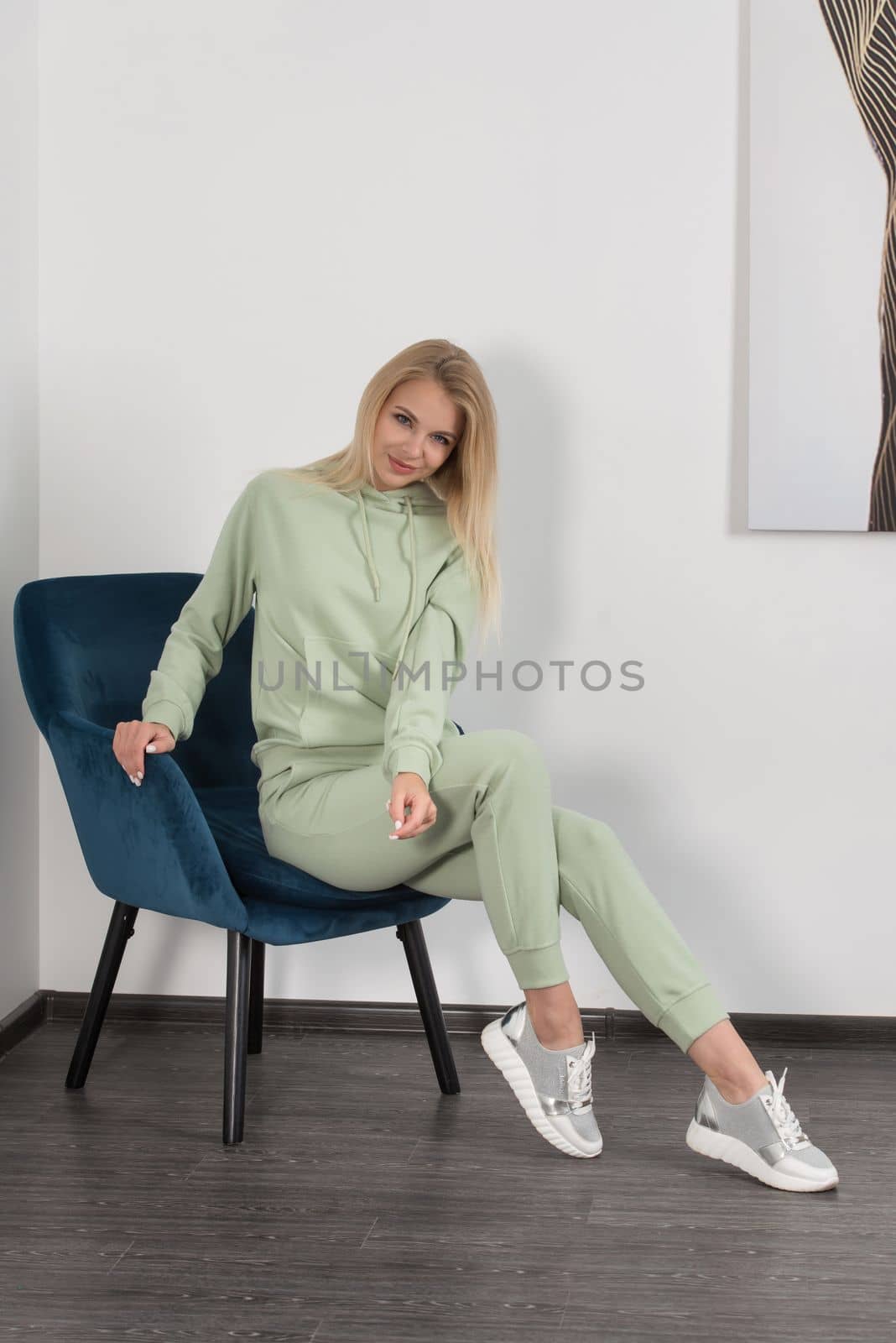 Stylish beautiful young blond woman in a light green tracksuit poses near a white wall in the room. Attractive girl model posing in a blue chair. by Ashtray25