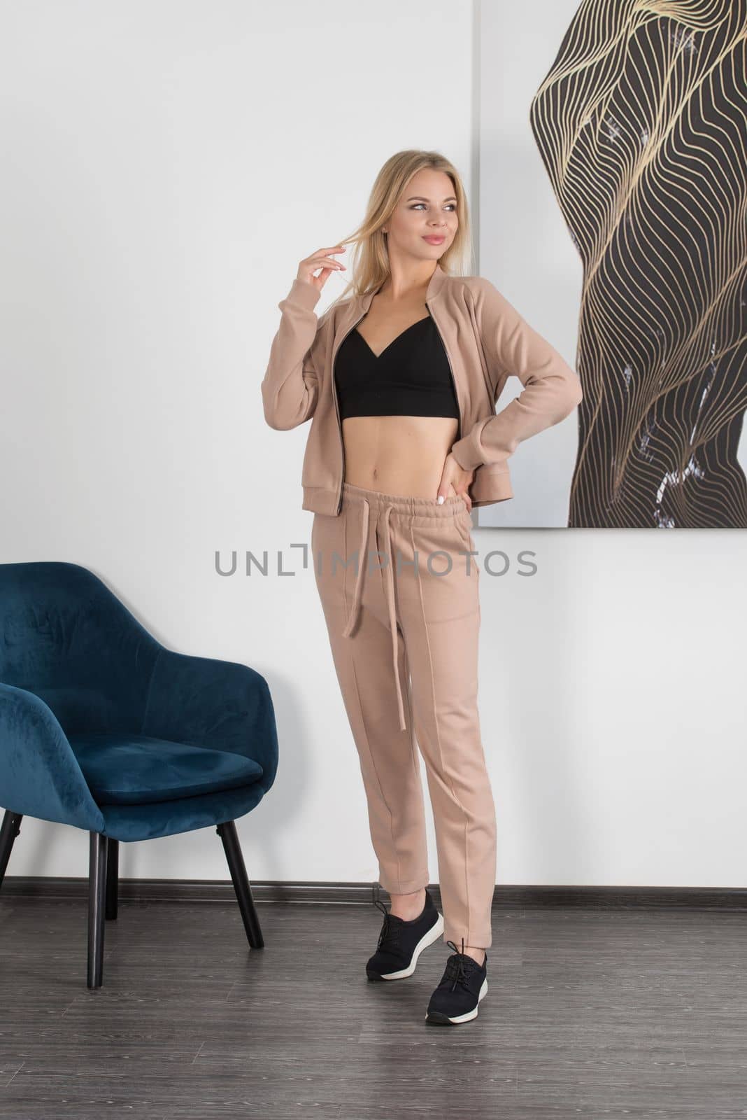 Stylish beautiful young blond woman in a biege tracksuit and black top poses near a white wall in the room. Fitness lady