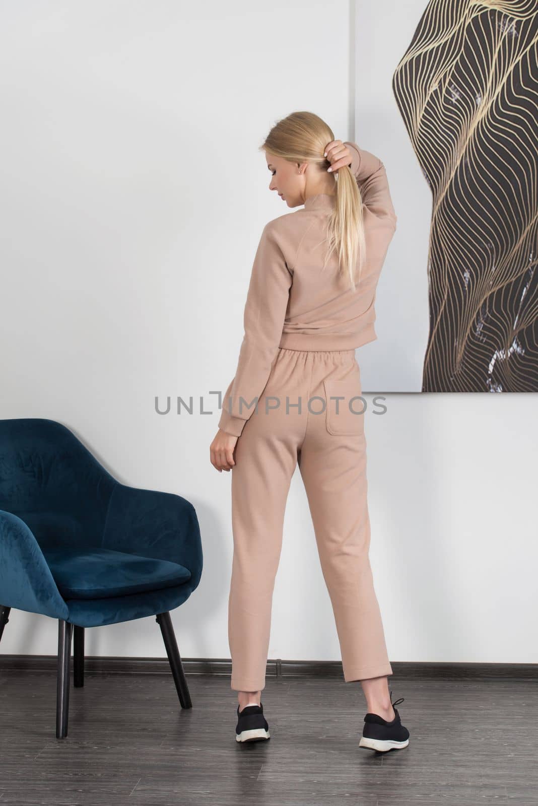 Stylish beautiful young blond woman in a biege tracksuit poses near a blue chair in the room. by Ashtray25