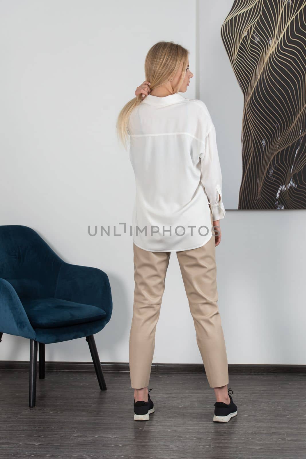 Stylish beautiful young blond woman in a tight biege pants and white shirt near a white wall in the room. Attractive girl model posing. by Ashtray25