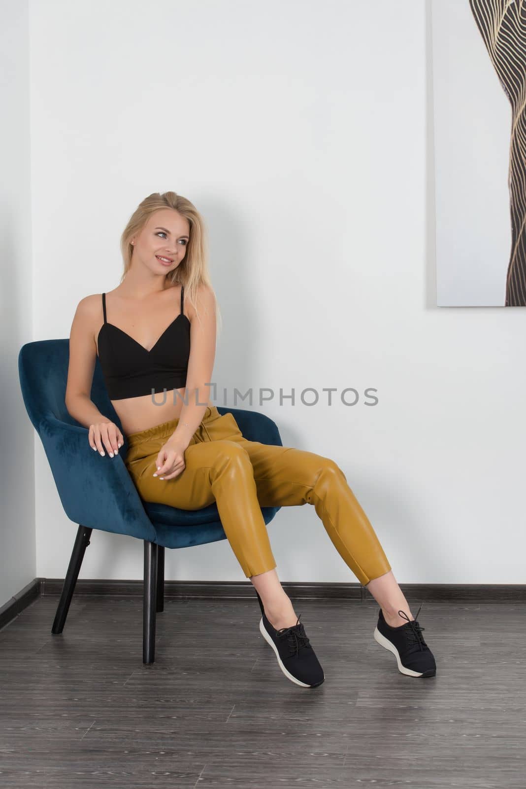 Stylish beautiful young blond woman in a tight yellow pants, black top and white shirt near blue chair in the room. Attractive girl model posing near blue chair. Fitness lady