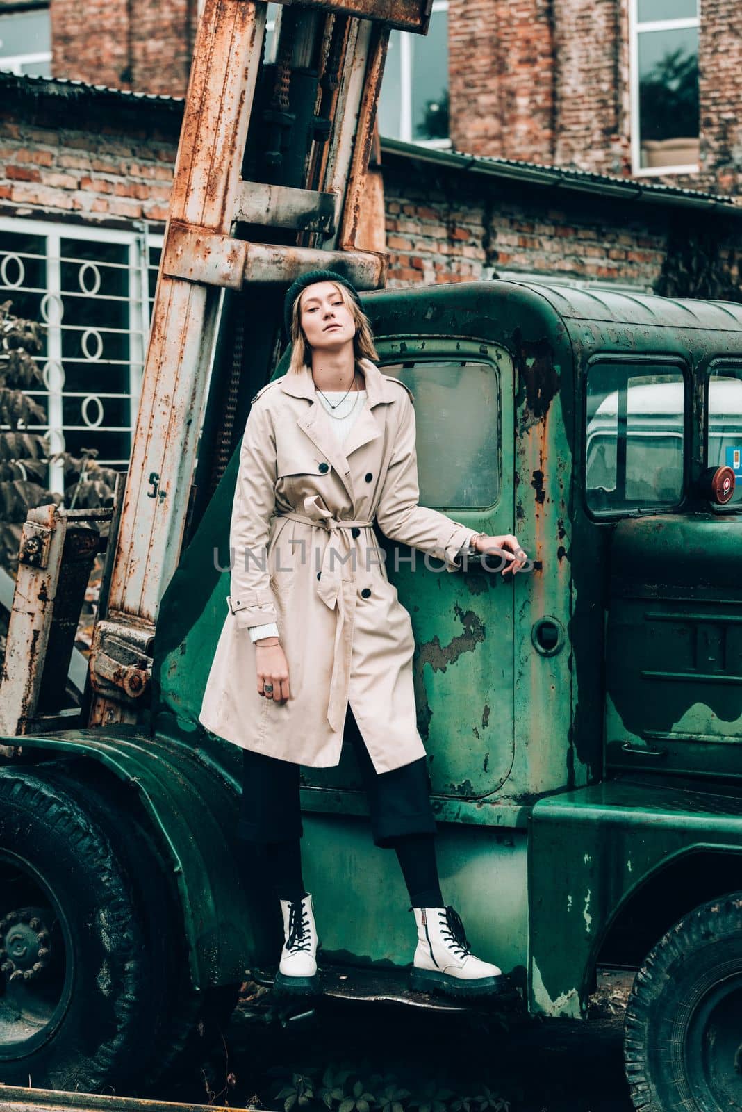 stylish blonde woman wearing long beige coat, white boots and black hat posing on the old green truck. Trendy casual outfit. Selective focus, grain by Ashtray25