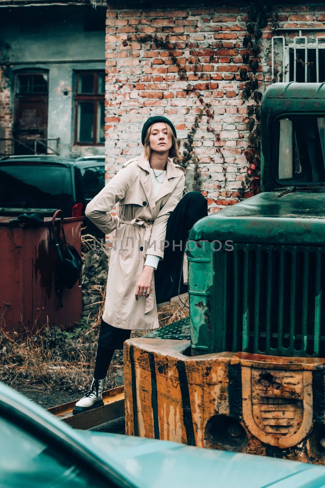 stylish blonde woman wearing long beige coat, white boots and black hat posing on the old green truck. Trendy casual outfit. Selective focus, grain by Ashtray25