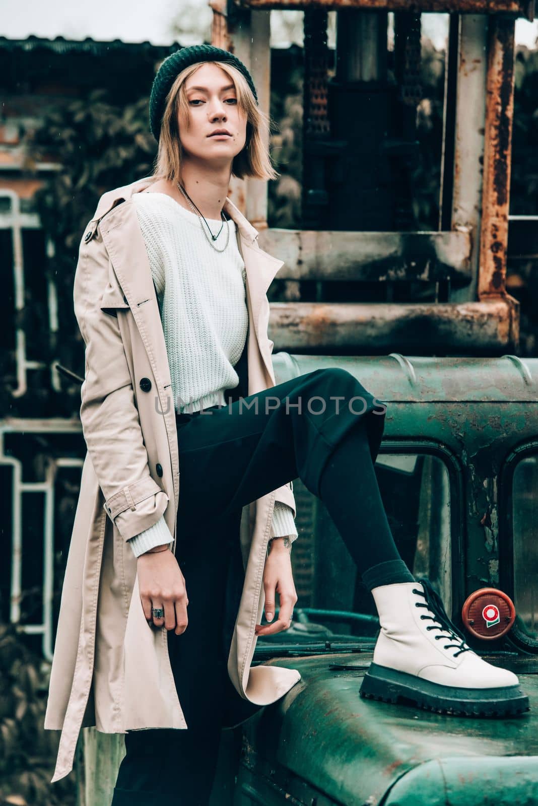 stylish blonde woman wearing long beige coat, white boots and black hat posing on the old green truck. Trendy casual outfit. Selective focus, grain by Ashtray25