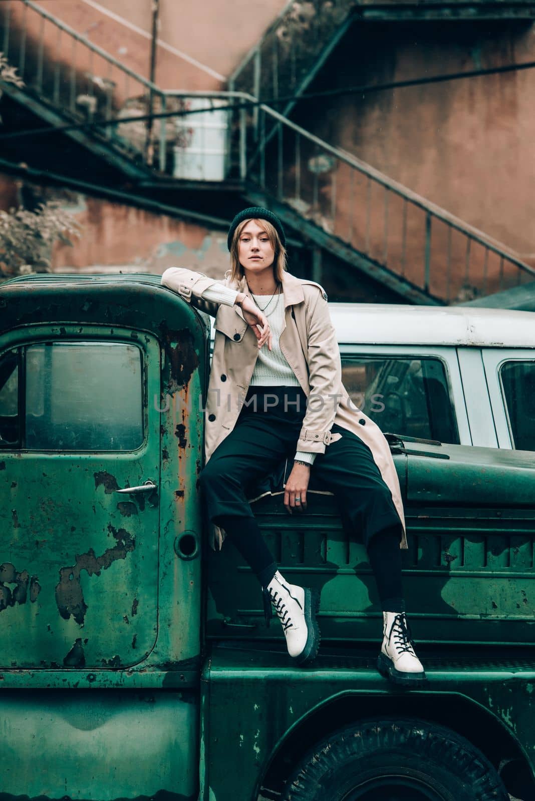 stylish blonde woman wearing long beige coat, white boots and black hat posing on the old green truck. Trendy casual outfit. Selective focus, grain by Ashtray25