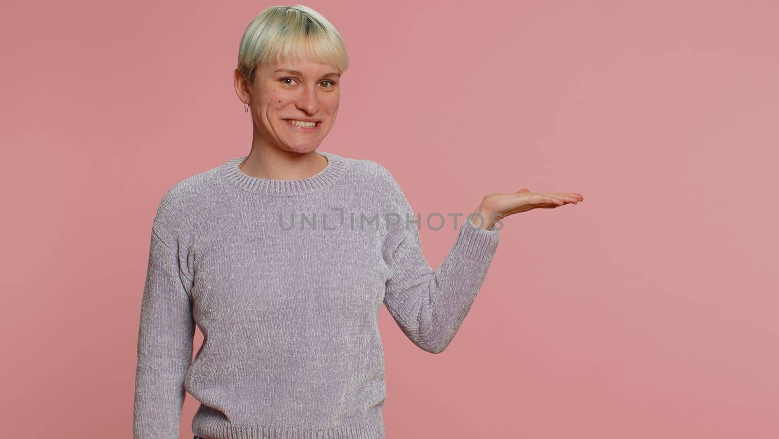 Lovely woman with short green hair showing thumbs up and pointing empty place, advertising area for commercial text, copy space for goods promotion. Young girl on pink background. Lgbt lesbian people