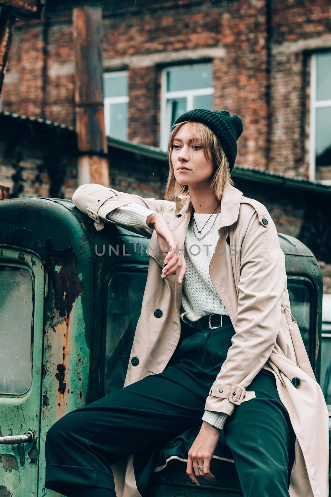 Beautiful young stylish blonde woman wearing long beige coat, white boots and black hat posing on the old green truck. Trendy casual outfit. Selective focus, grain. Street fashion. author's toning