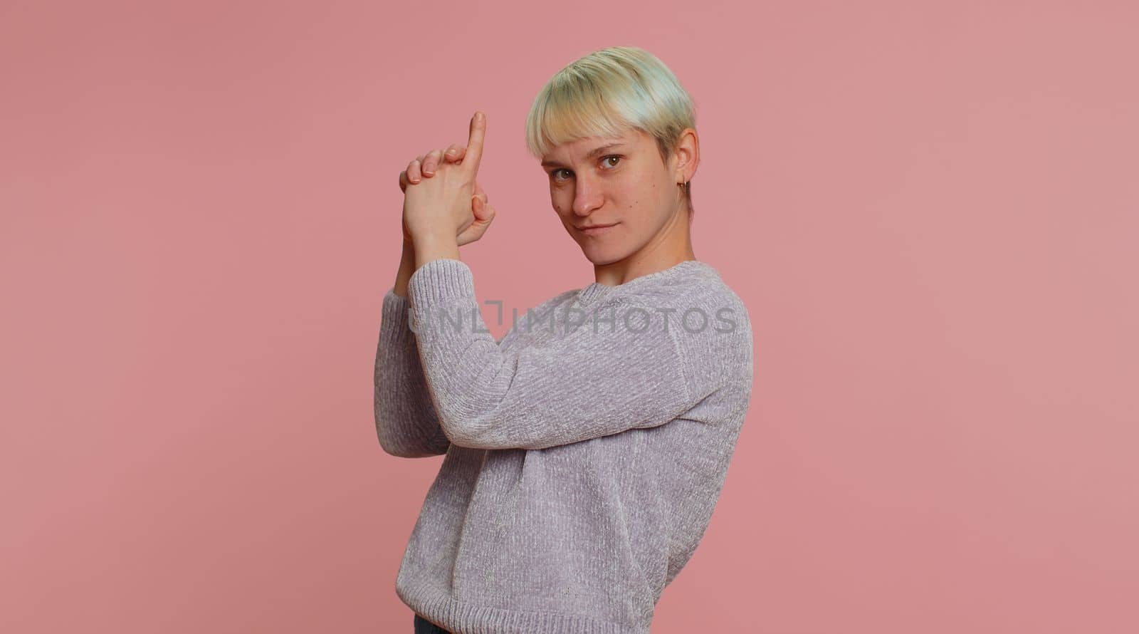 Woman with short hair pointing around with finger gun gesture, looking confident, making choice, shooting killing with hand pistol right on target. Girl on pink background. Lgbt gay lesbian people