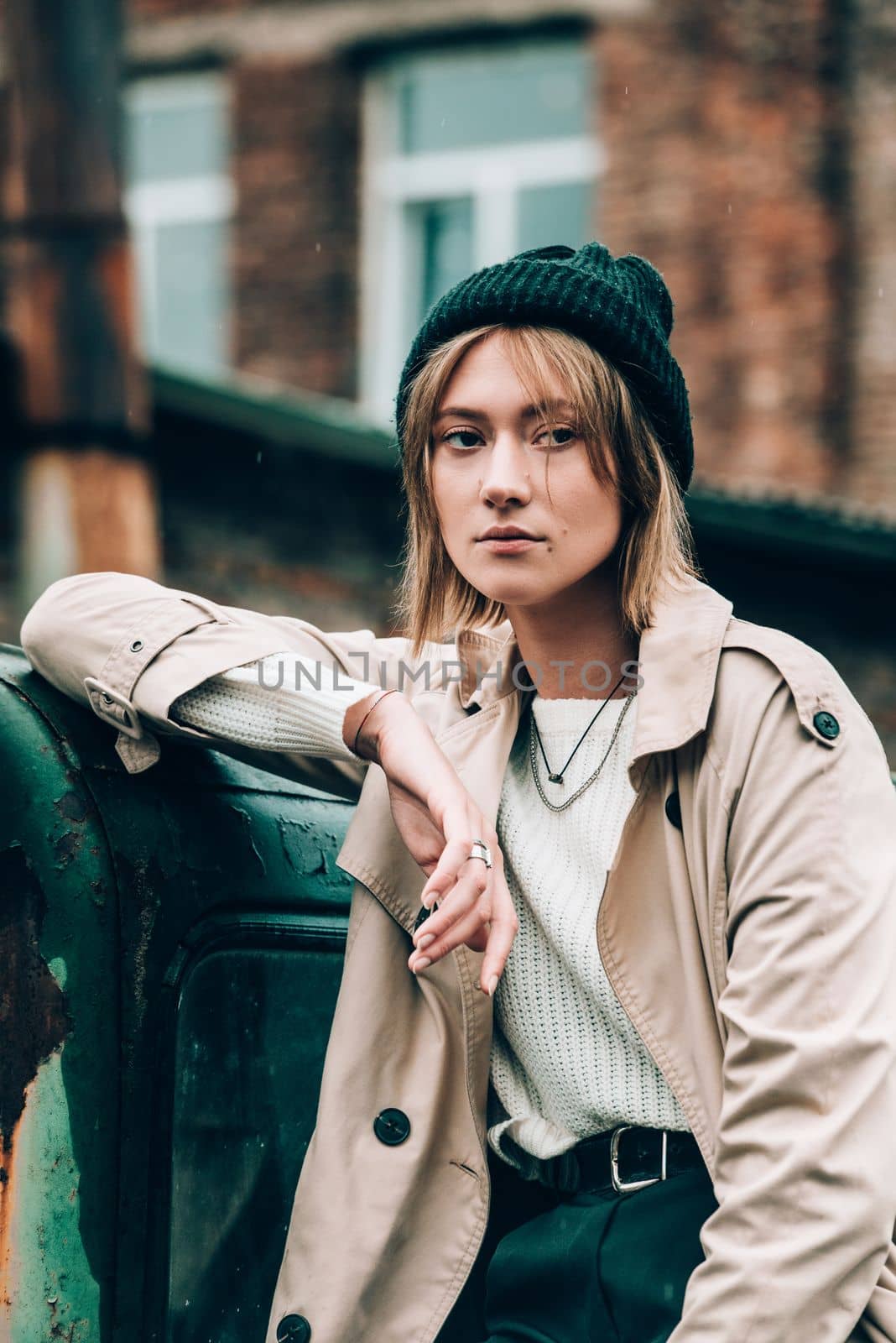 Beautiful young stylish blonde woman wearing long beige coat, white boots and black hat posing on the old green truck. Trendy casual outfit. Selective focus, grain. Street fashion. author's toning