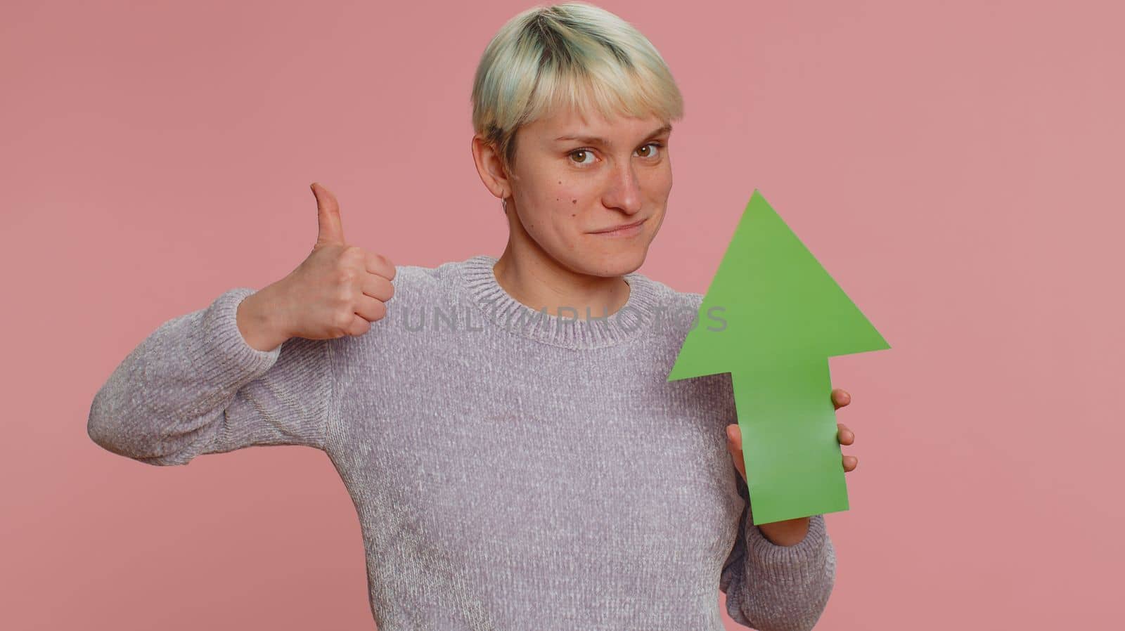 Young woman winner holding arrow sign pointing up, career growth and money exchange increase. Girl showing up indicating successful development progress on pink studio background. Lgbt gay lesbian