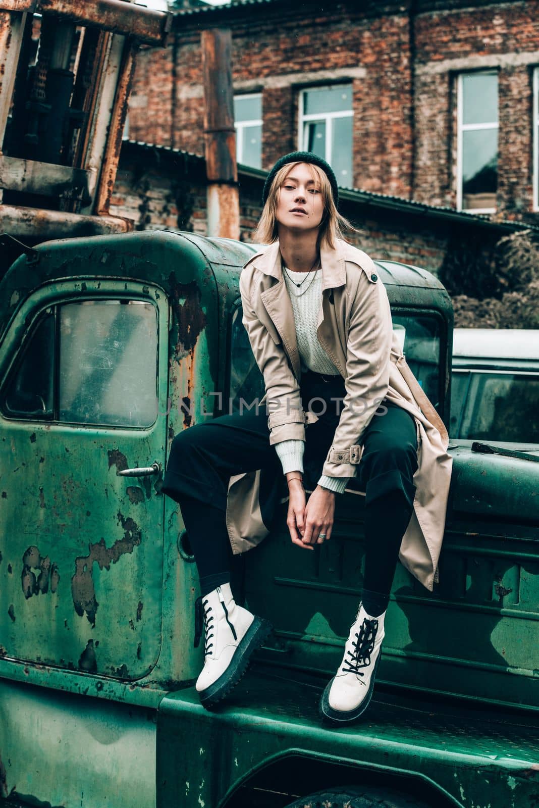 Beautiful young stylish blonde woman wearing long beige coat, white boots and black hat posing on the old green truck. Trendy casual outfit. Selective focus, grain. Street fashion. author's toning